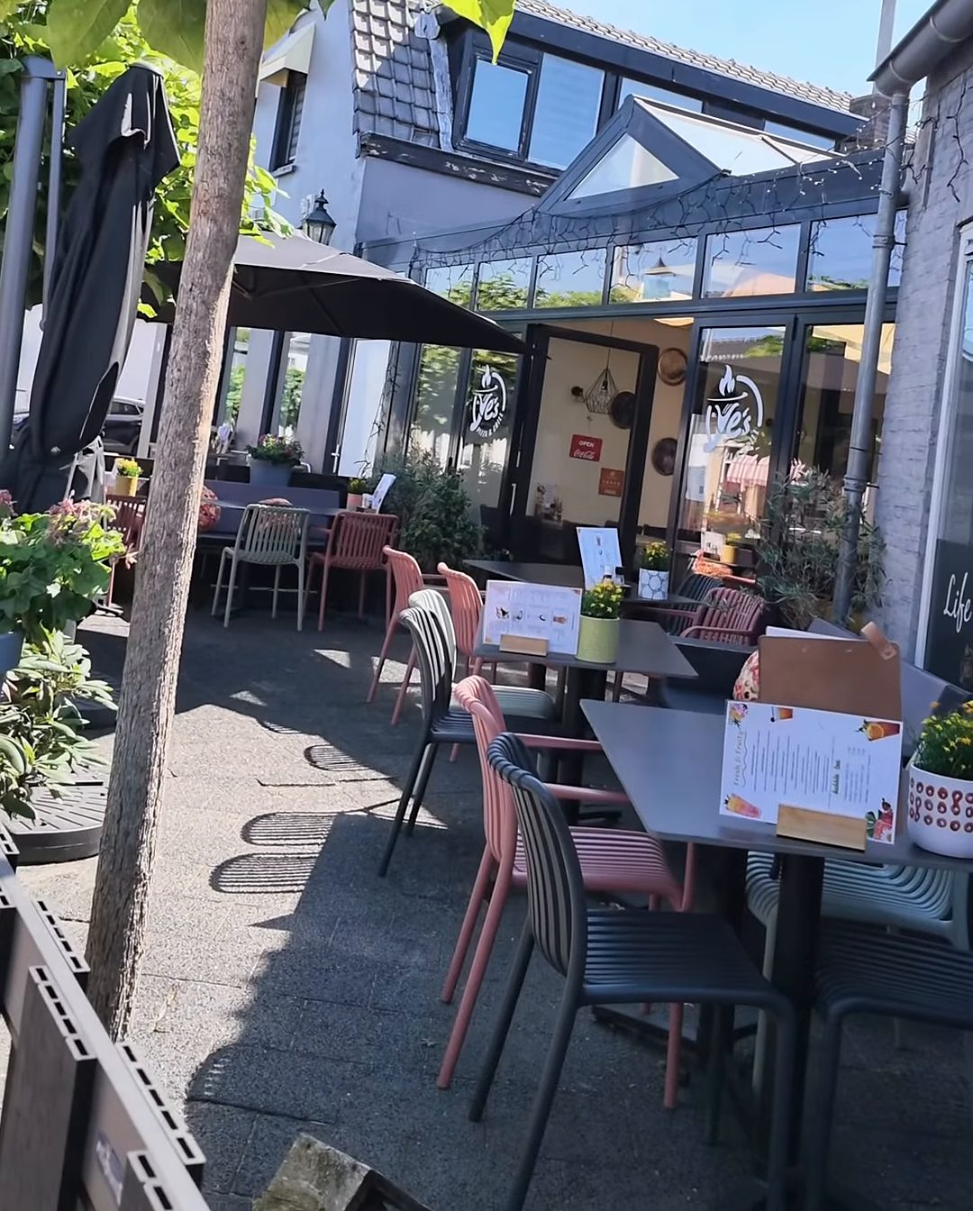Het terras van Ye's met bloembakken, bomen, parasols en roze stoelen